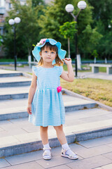 A little girl in a straw hat is standing on the steps in the park. A child in a beautiful blue dress.