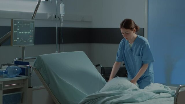 Caucasian nurse making hospital ward bed for use by sick person at modern medical facility unit. Emergency room with equipment, oxygen tube and wheelchair for intensive care treatment