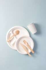 Obraz na płótnie Canvas Plastic-free disposable tableware on blue background, vertical. Paper plate, food containers, glass, wooden spoon, fork, knife. Single-use dinnerware non-plastic alternatives concept