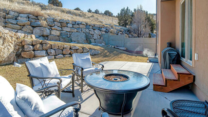 Pano Fire pit and paved patio at sunny backyard of house with stone retaining wall