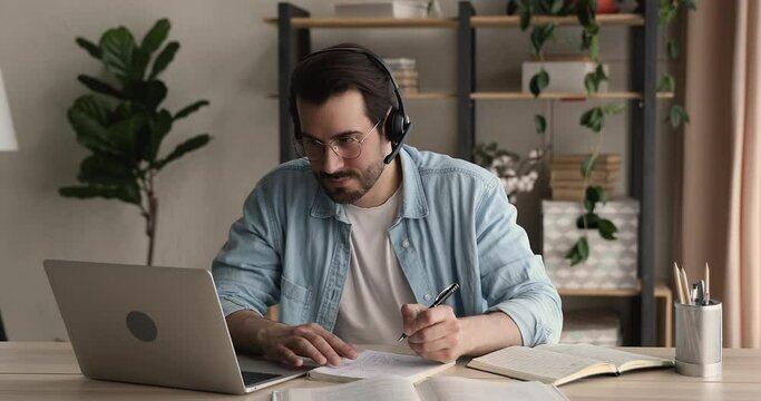 Focused young man in wireless headset with mic watching educational lecture online or holding web camera zoom video call, communicating distantly with teacher, improving professional knowledge.