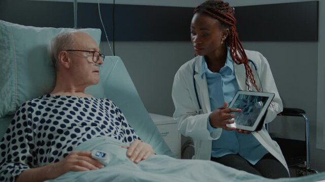 African american doctor showing xray on tablet to patient in hospital ward at medical facility. Sick elder man reads test results to start healing treatment for health and breathing problems