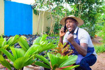 African male gardener, florist or horticulturist wearing an apron and a hat, making phone call and squatting as he works in a green and colorful flowers and plants garden