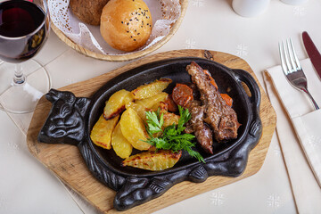 An iron plate with fried meat and potatoes, and herbs. A glass of wine and a basket of bread.
