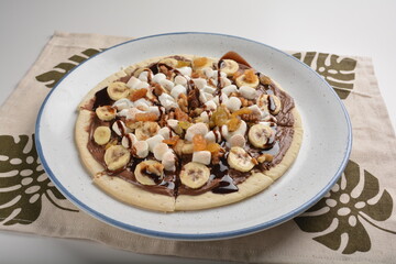 baked chocolate banana pizza with marshmallow and dry grape fruit dessert for kids in white background western halal menu