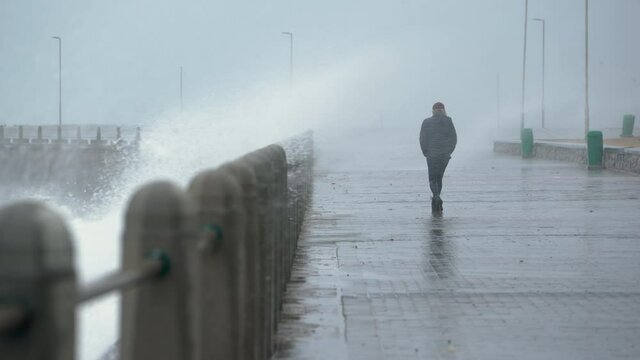 A lonely person walks in a raging storm along the promenade and gets soaked wet by the waves that crash over the seawall. Day.