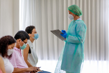 Young female citizen wears face mask in Coronavirus vaccinating queue line sitting checking her personal information from paper board in hand of full protective suit nurse who standing nearby