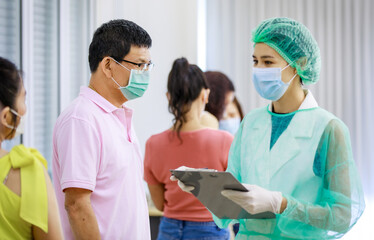 Young female nurse in full hazard protection uniform holding paper board asking personal information from senior male patient while others using application in smartphone in blurred background