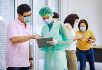 Fototapeta premium Young female nurse in full hazard protection uniform holding paper board asking personal information from senior male patient while others using application in smartphone in blurred background