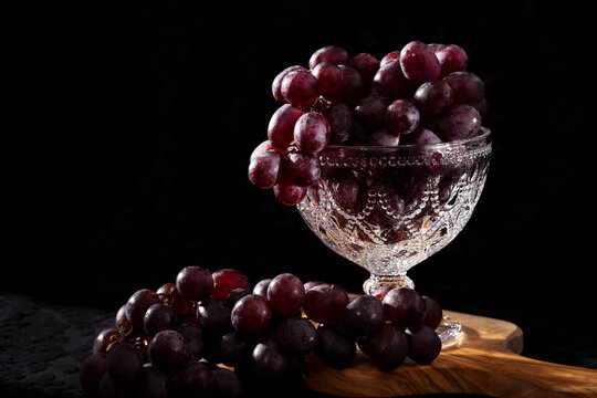Red, black grapes on a brown wooden cutting board. Dark, black background. Morning light, ray shining on the grapes and crystal. retro, bohemic style.