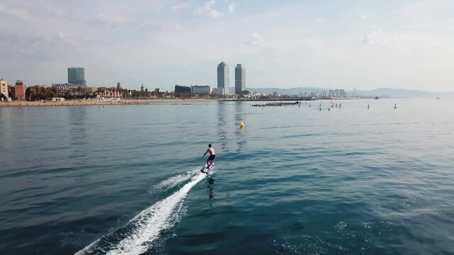 Aerial follow shot of fast surfer riding electric surfboard in front of iconic Barcelona skyline on a sunny day