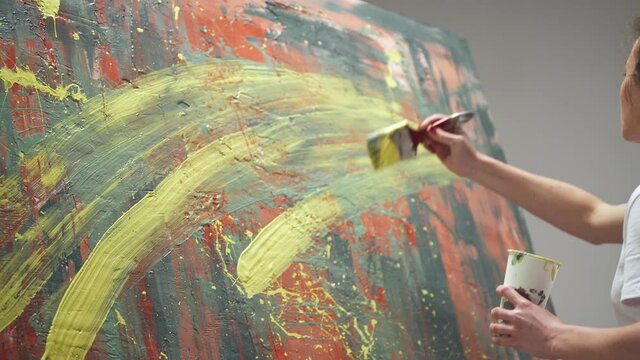 Young woman artist draws with a brush on a large canvas, a talented artist draws a color red abstraction, splashing paint on canvas, back view.