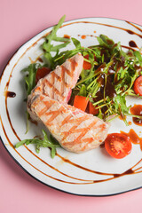 Close up still life of a tuna steak with fresh vegetables and arugula with seasoning over pastel pink background.