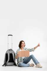 Young woman with cardboard and suitcase hitchhiking on light background