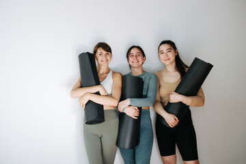Trio of joyful young women posing with yoga mats in hands, smiling. All three are wearing sportswear. Over white wall.