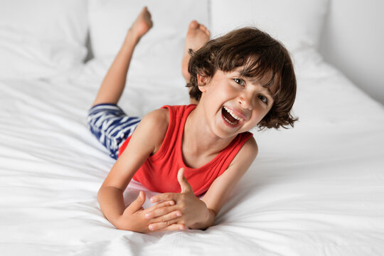 Laughing young boy lying on white bed resting on elbows with crossed hands