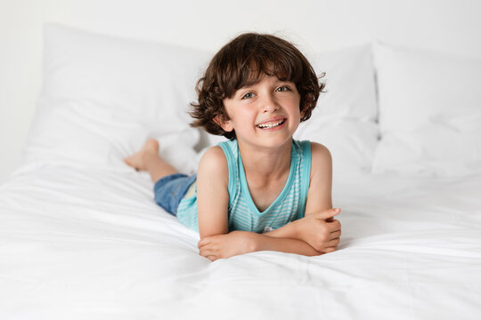 Smiling young boy lying on white bed resting on elbows