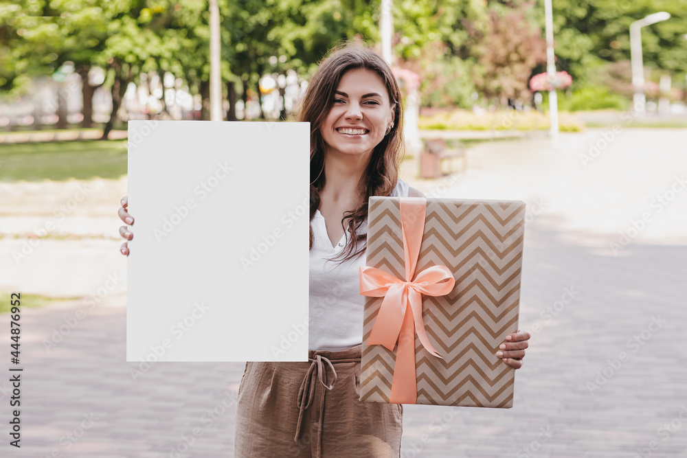 Wall mural young woman holding a white poster in her hands and packaging with a bow and smiling against the bac