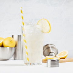 Homemade traditional refreshing lemonade preparation - cold citrus drink with ice, straw in glass with ingredients, silver crokery in modern white kitchen interior, square.
