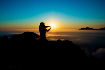 Silhouette of a woman catching the sun with her hands, on top of a mountain. sunset and cloud sky background.
