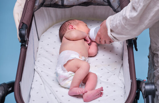 Mother hands give a bottle of milk to a newborn baby in a stroller, blue background