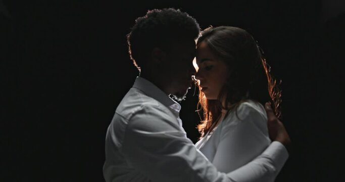 Young elegant couple of different nationalities in tender passion, embrace each other, man cares, comforts his woman, gently touches her hair, shows love, shot on black background in twilight