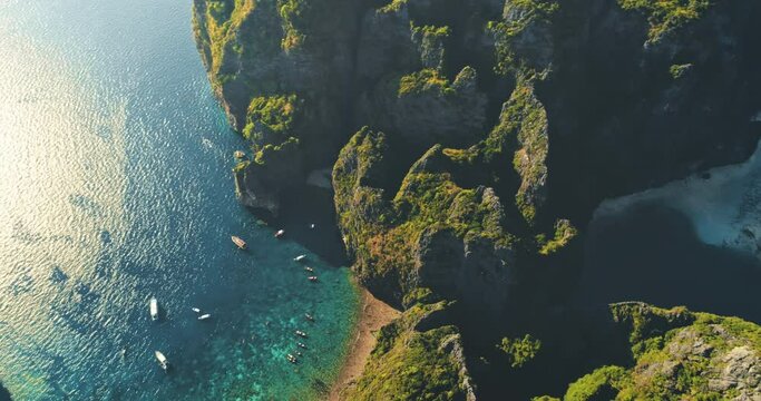 Bright sun shines on rocks and azure sea water, aerial view. Green grass on mountains near blue ocean. Drone flying over boats in bay. Beautiful wild landscape Phi Phi island, Thailand, slow motion