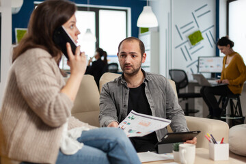 Employee taking at smartphone with business partner sitting on couch looking at documents. Woman discussing on phone while multiethnic coworkers planning financial project working in open plan office