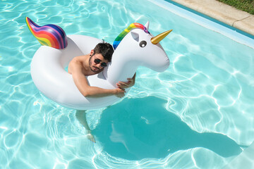 Young man wearing sunglasses hugging a big unicorn inflatable ring in a swimming pool. Summer concept.