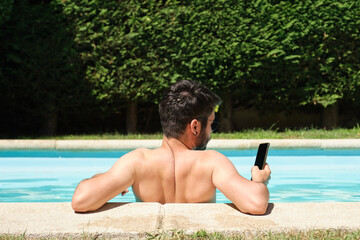 Young man wearing sunglasses using a smartphone backwards in a swimming pool. Summer concept.