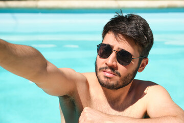 Young man wearing sunglasses taking a selfie with a smartphone in a swimming pool. Summer concept.