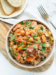 Stewed buckwheat with meat, chickpeas and vegetables, decorated with fresh herbs, served in a round bowl on a light background top view, next to a fork and two slices of bread. A delicious, hot lunch.