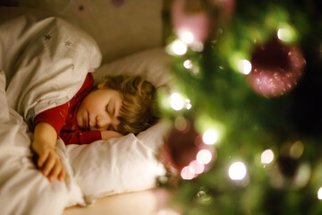 Little cute toddler girl sleeping under Christmas tree and dreaming of Santa at home, indoors. Traditional Christian festival. Happy kid child waiting for gifts on xmas. Cozy soft light