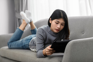 Attractive young women lying using a digital tablet on the sofa in the living room, modern and new normal lifestyle relaxing at home.