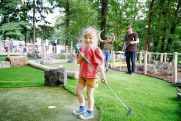 Cute preschool girl playing mini golf with family. Happy toddler child having fun with outdoor activity. Summer sport for children and adults, outdoors. Family vacations or resort. - obrazy, fototapety, plakaty