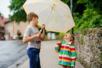 School kid boy picks up the little sister, preschool girl from kindergarten. Two happy children with umbrella playing during rain and having fun on bad weather rainy day. Siblings in love.
