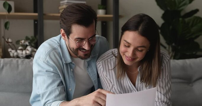Smiling young family couple reading paper letter with positive news, discussing bank loan mortgage conditions, job offer, business agreement, learning contract, analyzing information together at home.