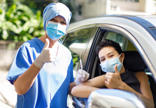 Female Patient Sit In Car And Caucasian Doctor Wear Face Mask Hold Covid 19 Syringe Needle Vaccine Stand Hold Thumb Up Together Look At Camera In Drive Thru Vaccination Queue