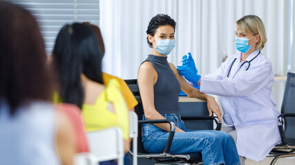 Obraz premium Cute female wears face mask sit look at camera while Caucasian woman doctor in white lab coat blue gloves and stethoscope injecting shot vaccine on shoulder when other patients wait in queue line