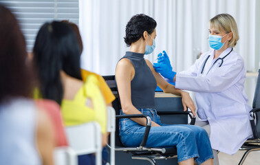 Obraz premium Cute female wears face mask sit look at camera while Caucasian woman doctor in white lab coat blue gloves and stethoscope injecting shot vaccine on shoulder when other patients wait in queue line
