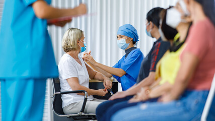 Obraz premium Asian female doctor wears face mask and hospital uniform injecting coronavirus vaccine shot to Caucasian senior woman shoulder while other multi ethnics patients looking in blurred foreground queue