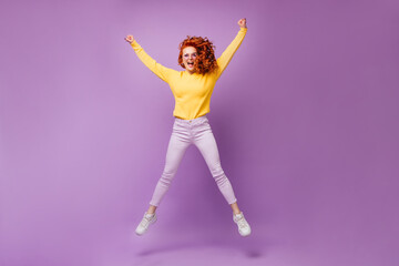 Fototapeta na wymiar Red-haired girl in glasses and yellow sweater jumping on purple background