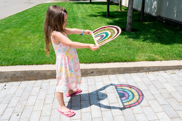 Cute child girl in tie dye dress playing with rainbow suncatcher outside. Fun crafting ideas for kids from recyclable items. Creative play with shadows. DIY concept