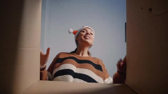 Caucasian woman with santa hat opening present box for surprise at home. Young festive adult smiling looking inside package. Person receiving christmas gift from friends and relatives. View from