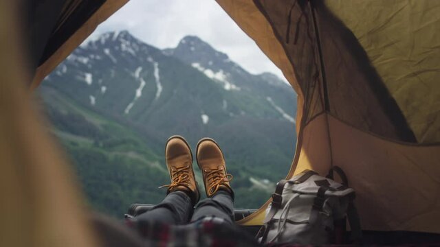 Women's legs in yellow hiking boots lie in a yellow tent. The traveler is resting, admiring the mountains after climbing. Yellow boots against the background of big mountains. Rest in the camp.
