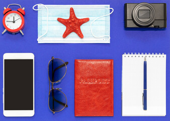 A set of necessary accessories for a trip on a blue background: a tourist's passport , a camera, a phone, a notebook, a protective mask and sunglasses. The concept of tourism. Flat layout.