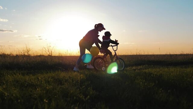 Childhood Bike Concept. Mother Teaching Son To Ride Bicycle. Happy Cute Boy Learn To Riding A Bike In Park At Sunset Time. Young Mom Teaching Son To Ride Bike First Time On Countryside Rural Road.