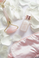 Face skin care products set. Face roller, serum, sleep mask with rose petals on white background. Flat lay, top view.