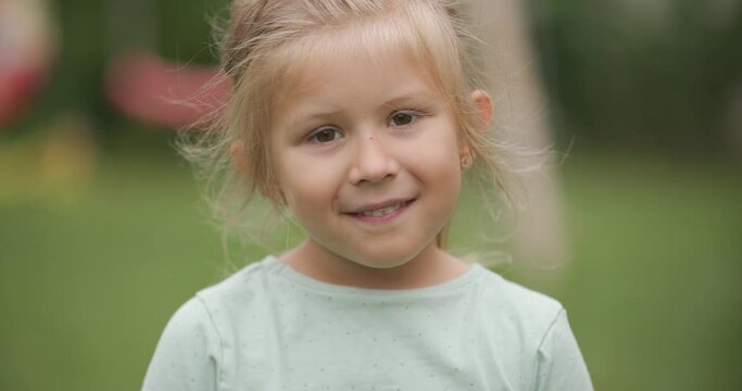 cute blonde kid with innocent eyes being timid, looking at camera and smiling while playing outside in backyard