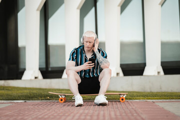 blond man sitting on skateboard while listening to music with headphones on the street - Powered by Adobe